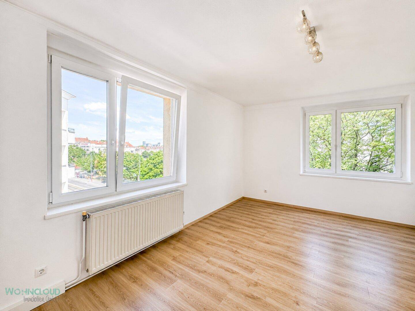 Helles, leeres Zimmer mit zwei großen Fenstern und Blick ins Grüne.