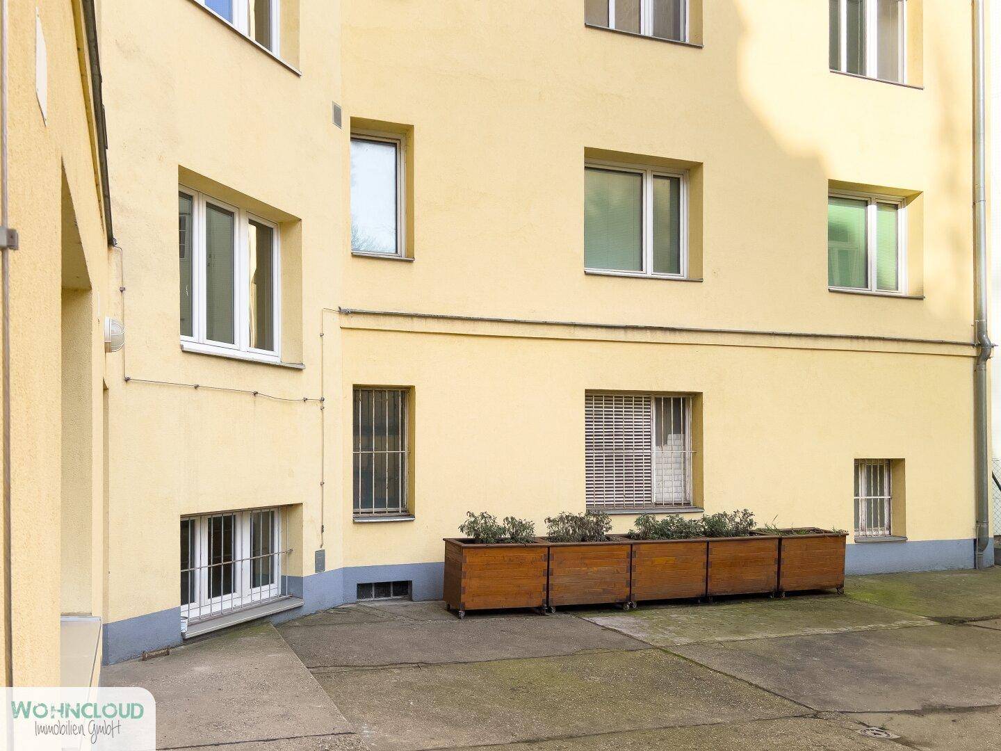Hinterhofbereich eines Gebäudes mit gelber Fassade und Holzpflanzkübeln.