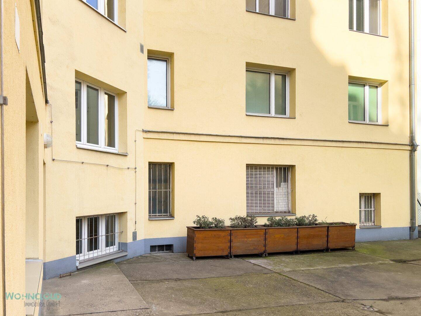 Gelbe Gebäudefassade mit mehreren Fenstern und einem begrünten Innenhofbereich.