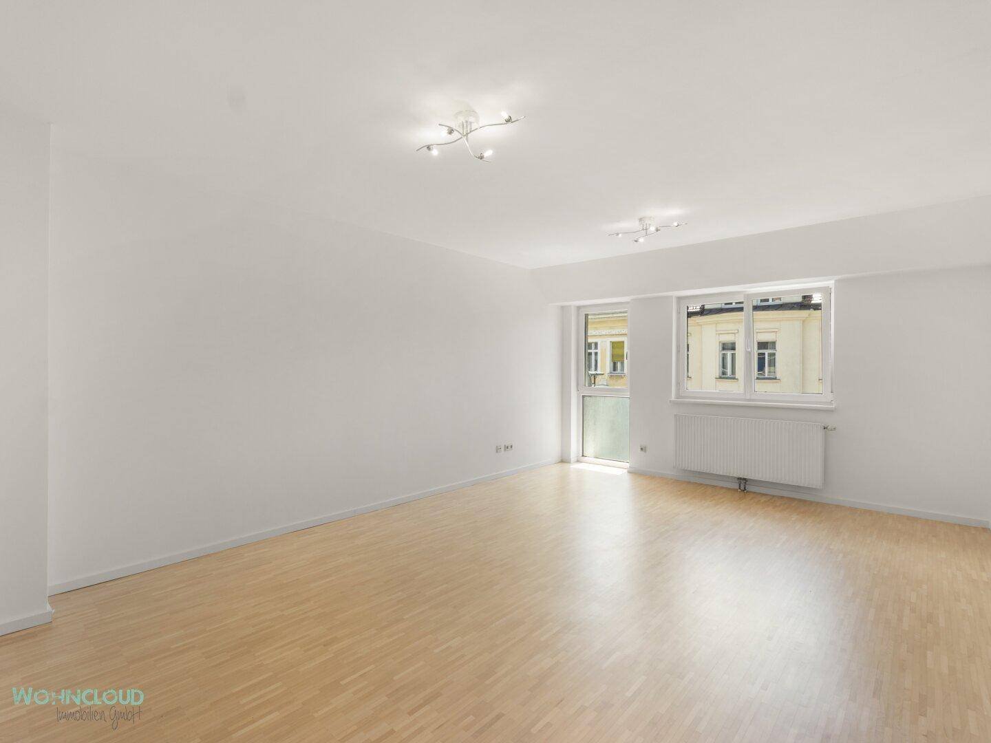 Geräumiges, leeres Wohnzimmer mit hellem Parkettboden und großen Fenstern.