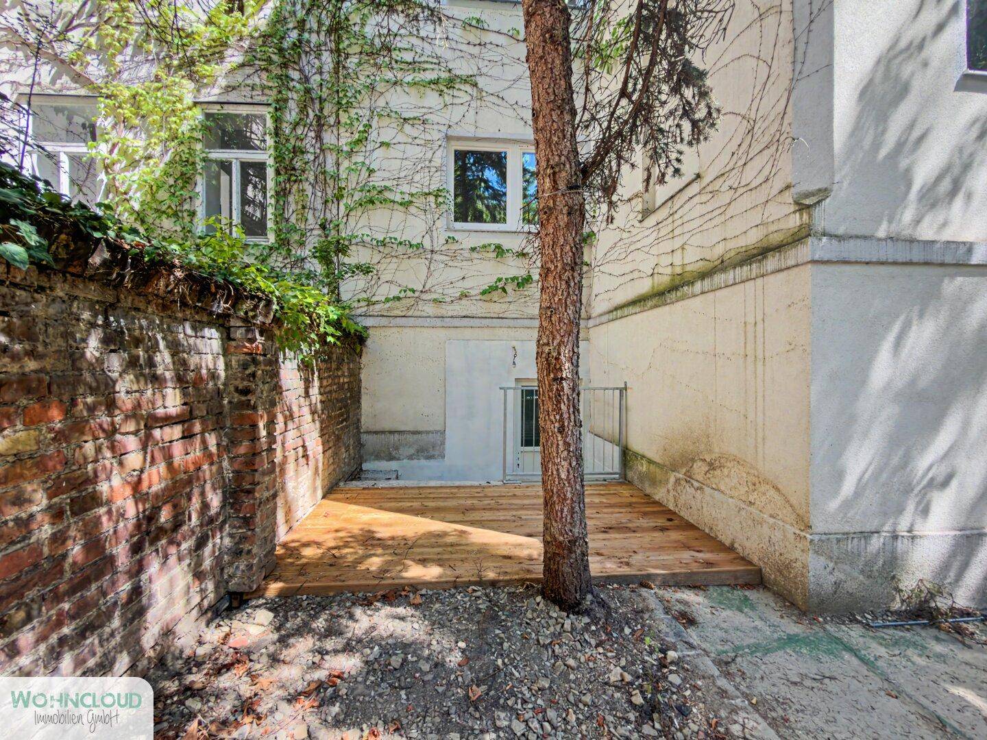 Unmöblierter Außenbereich mit Holzterrasse, alter Ziegelmauer und Baumbestand, bereit zur Gestaltung.