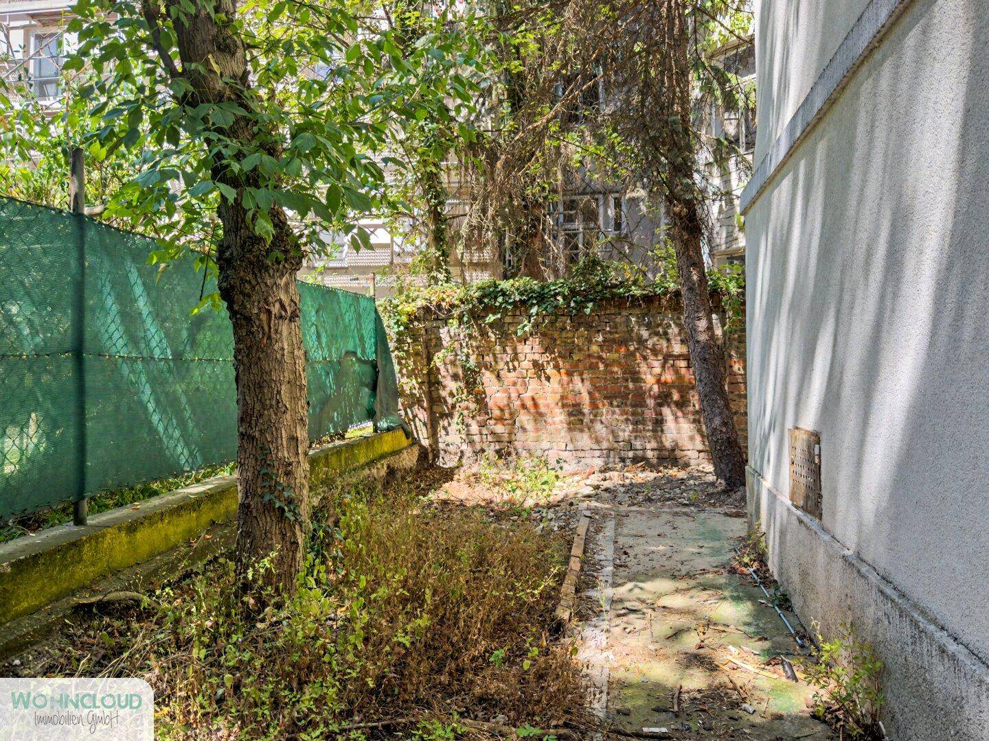 Naturbelassener Gartenbereich mit altem Baumbestand, Ziegelmauer und unbefestigtem Weg.