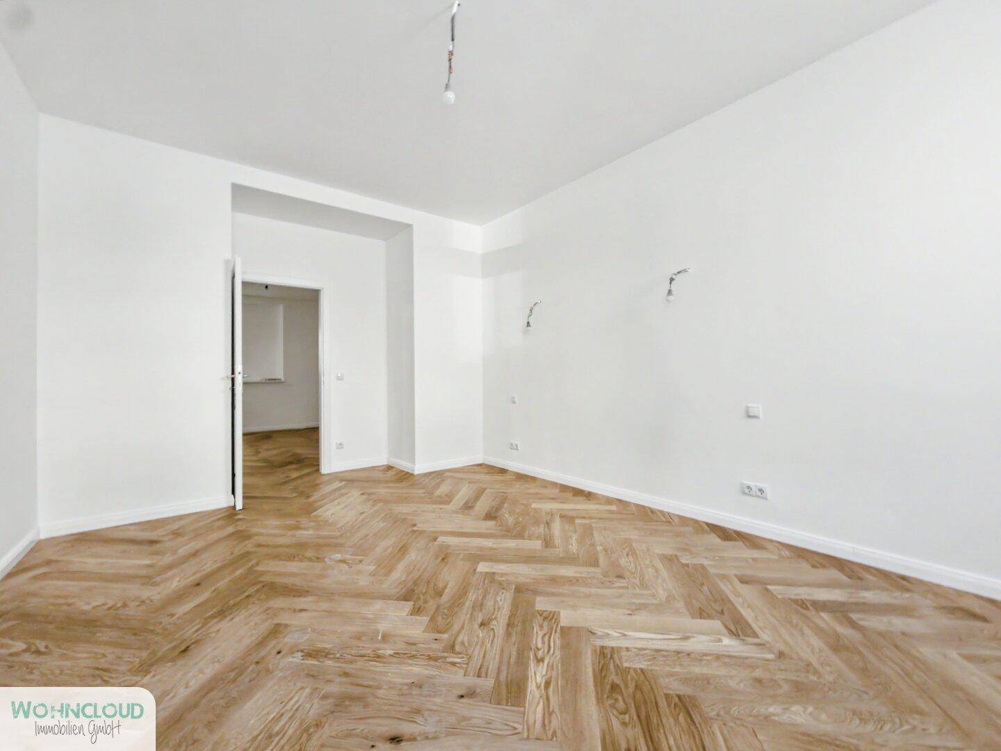 Geräumiges, unmöbliertes Zimmer mit hellem Fischgrätparkett und weißen Wänden, vielseitig nutzbar.