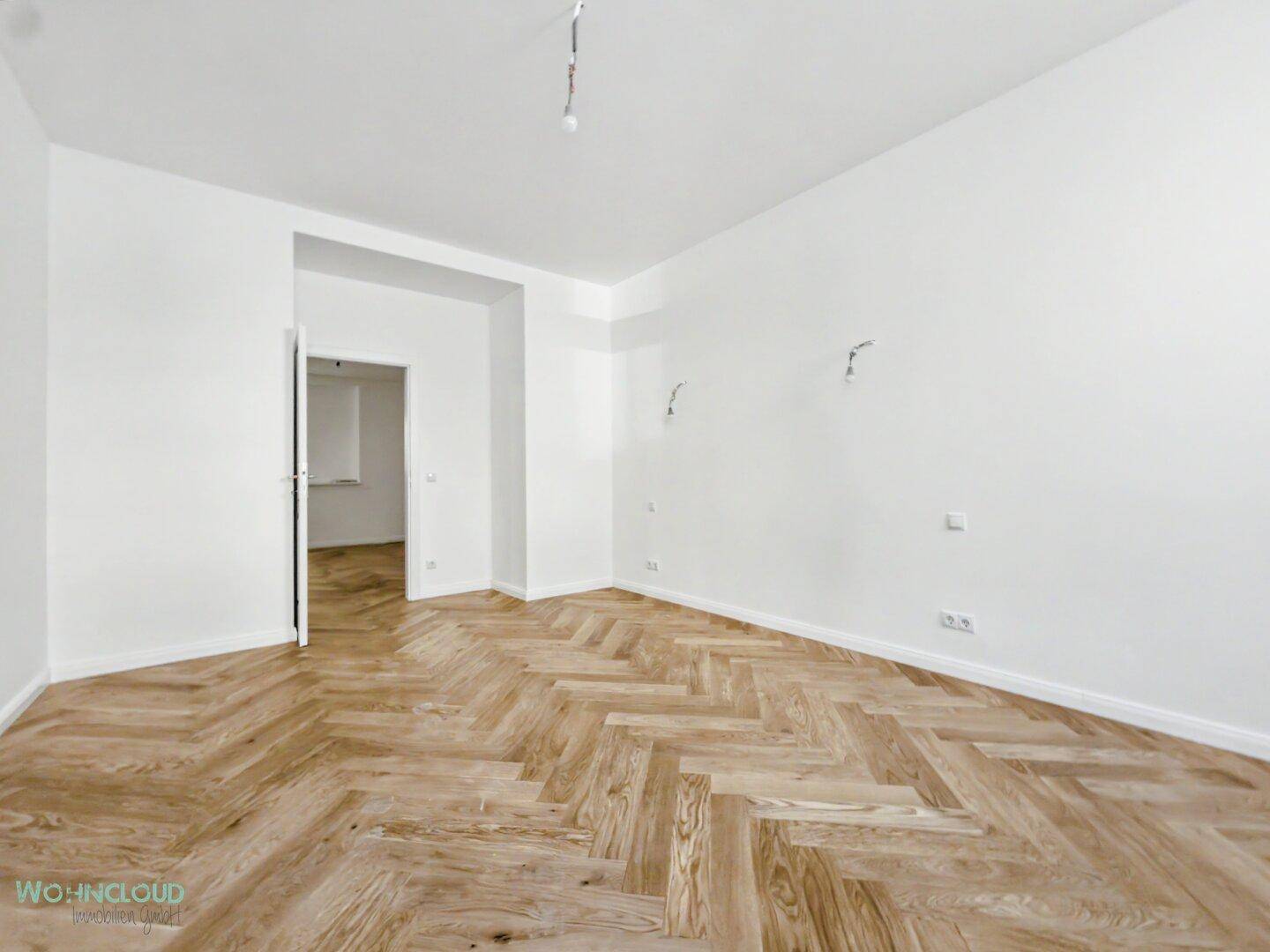 Geräumiges, leeres Zimmer mit Fischgrätparkett und weißen Wänden, vielseitig nutzbar.