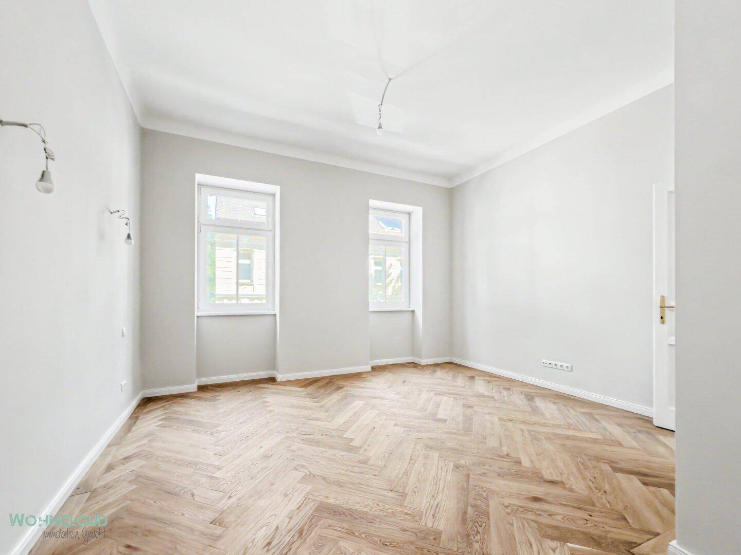 Leeres Zimmer mit Fischgrätparkett und zwei Fenstern, ideal als Schlaf- oder Arbeitszimmer.