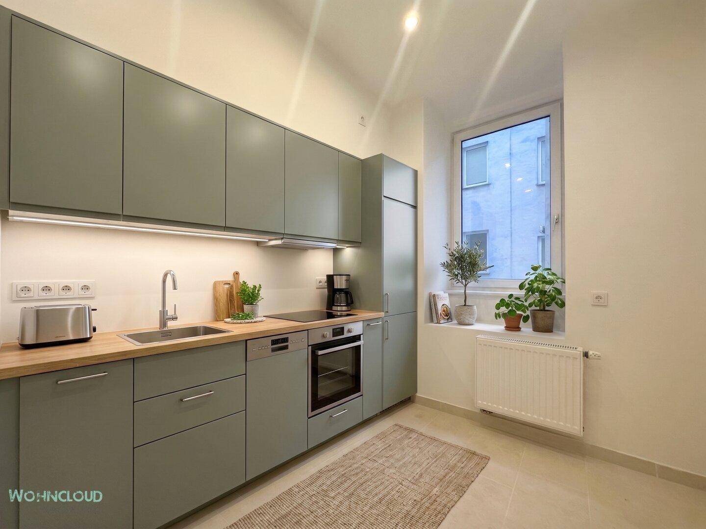 Moderne Küche mit mattgrünen Schränken, Holzarbeitsplatte und einem Fenster, das Tageslicht spendet.