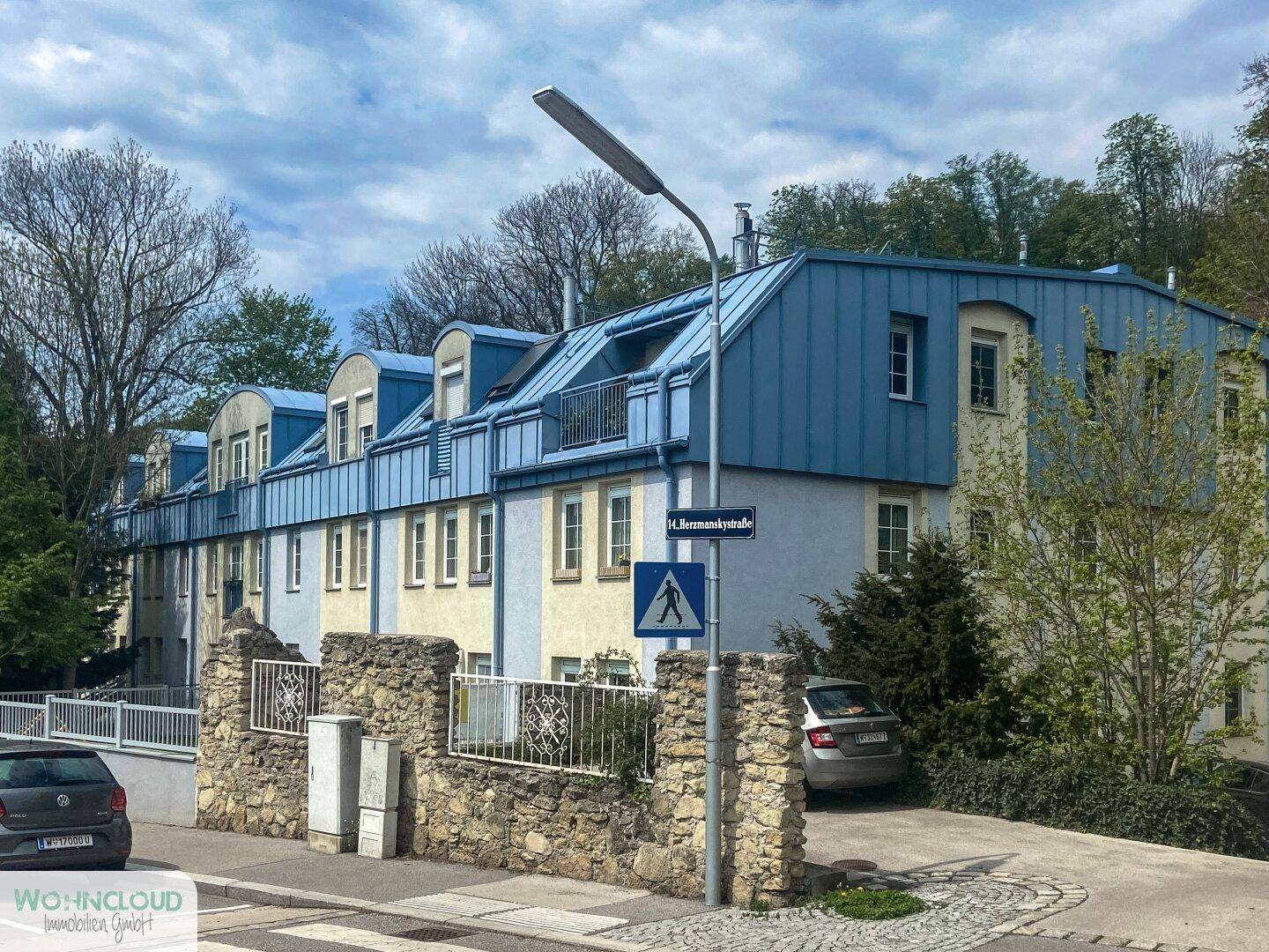 Mehrfamilienhaus mit markantem blauen Mansarddach und heller Fassade an einer Straße.