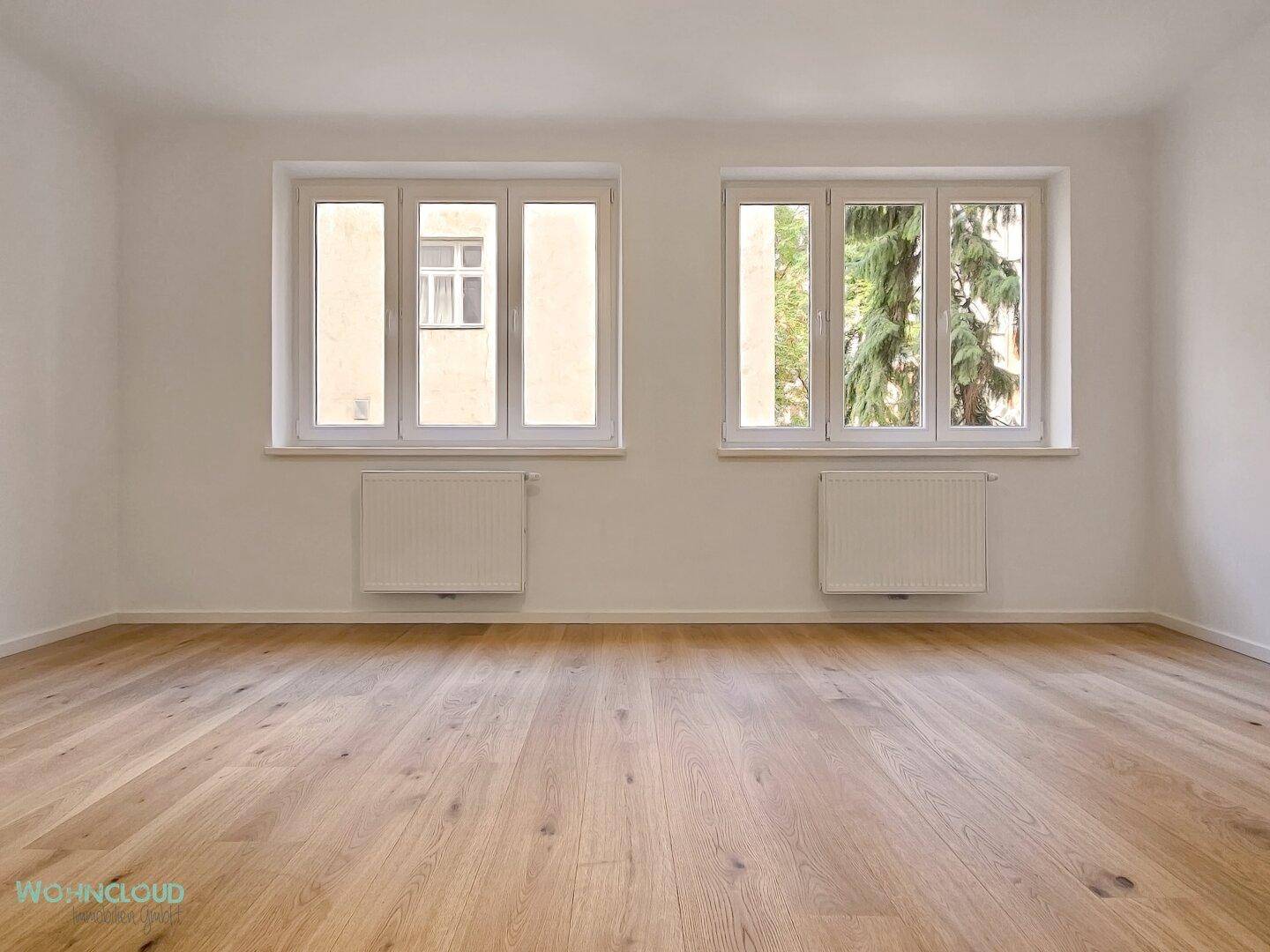 Geräumiger, leerer Raum mit hellem Holzparkett, weißen Wänden und zwei großen Fenstern mit Grünblick.