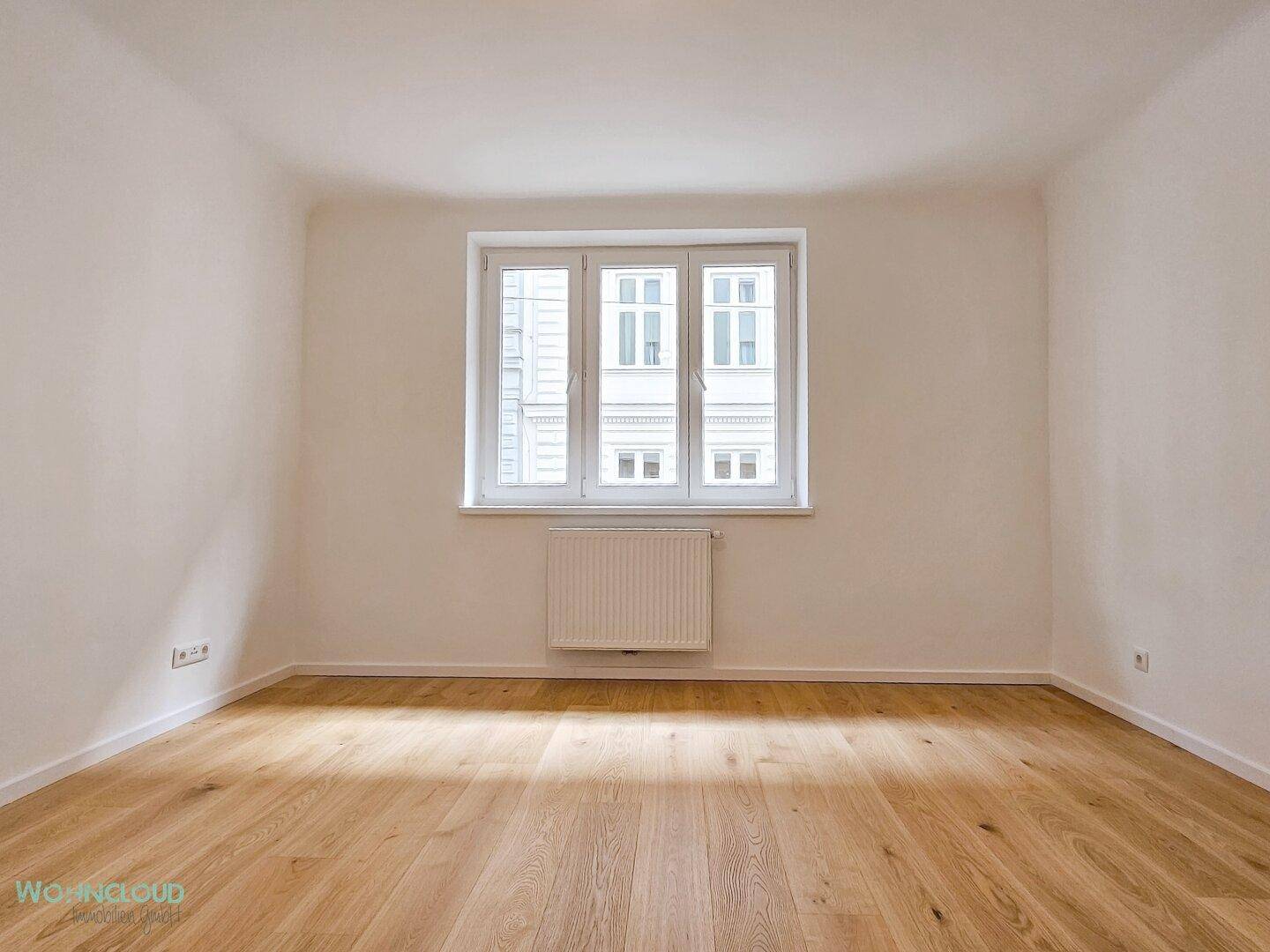 Helles Schlafzimmer mit neuem Holzparkettboden, weißen Wänden und einem großen Fenster zur Straße.
