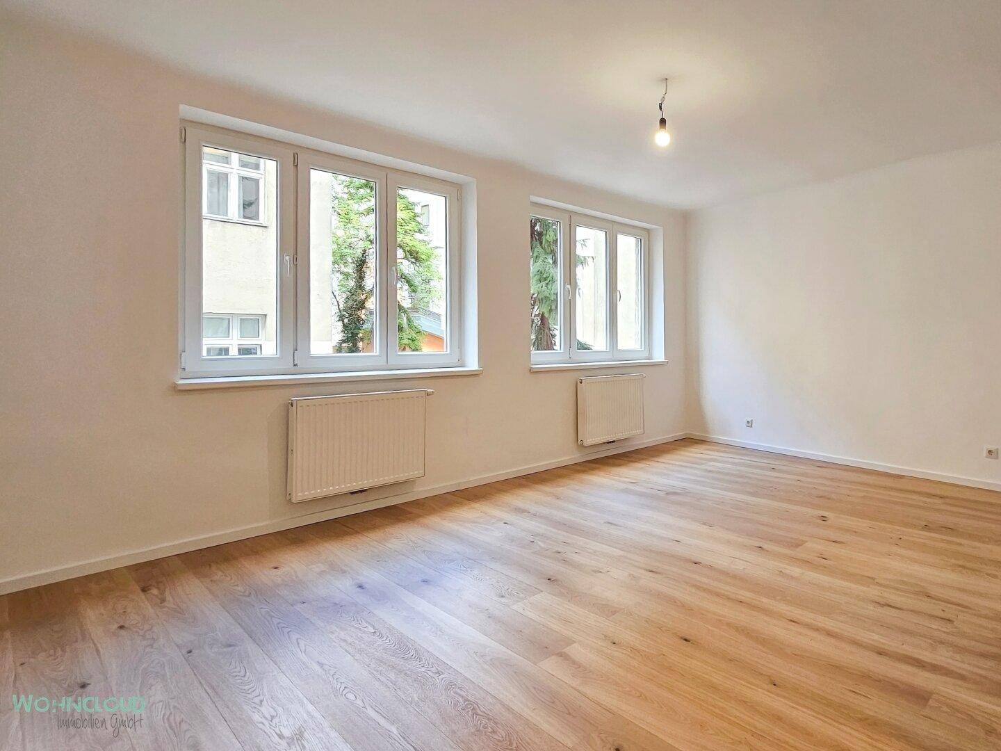 Lichtdurchfluteter Raum mit neuem Holzparkettboden, weißen Wänden und zwei Fenstern mit Blick ins Grüne.