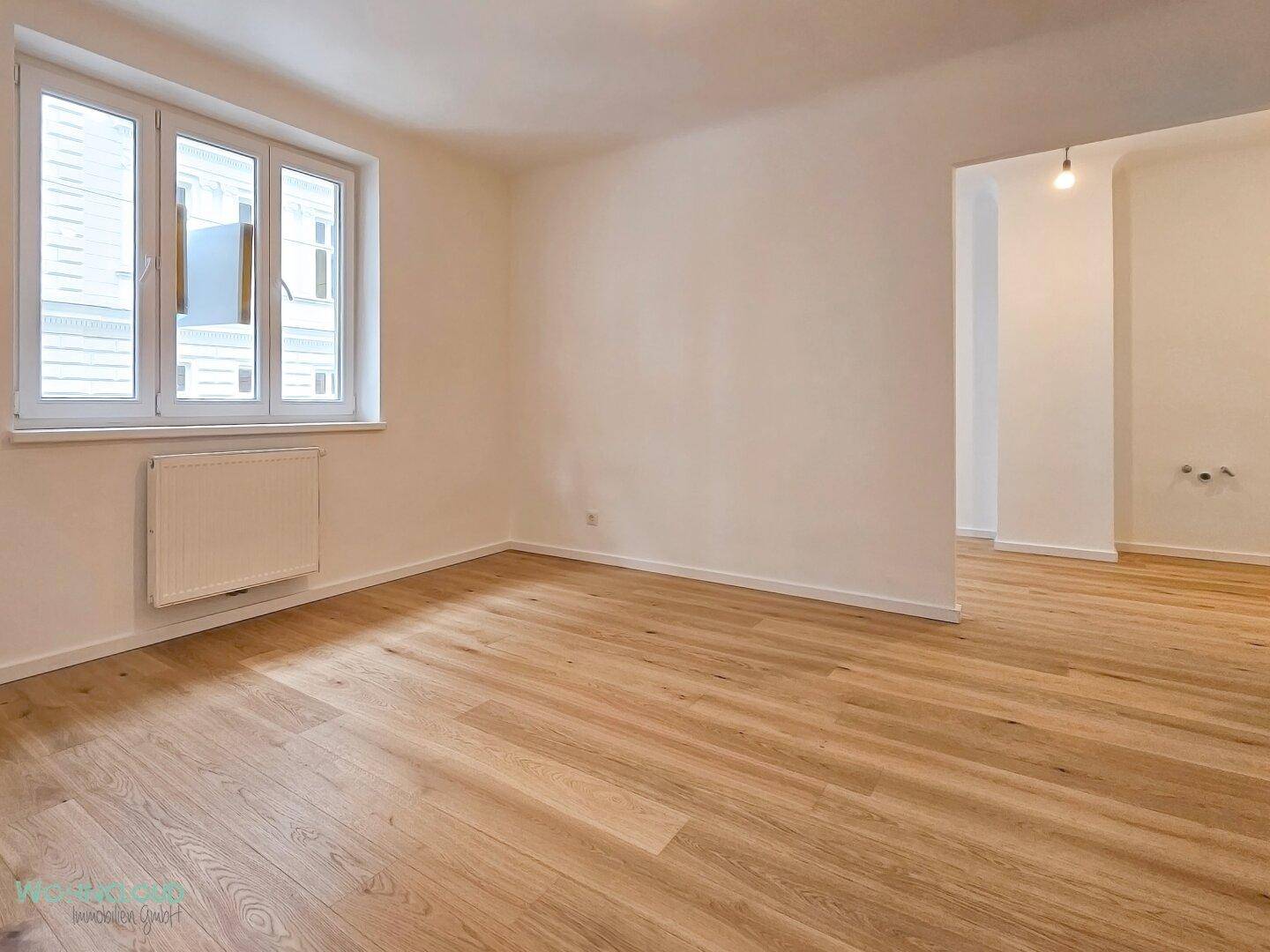 Geräumiger, heller Wohnraum mit Holzparkettboden, weißen Wänden und Fenster, offen zu einem weiteren Bereich.