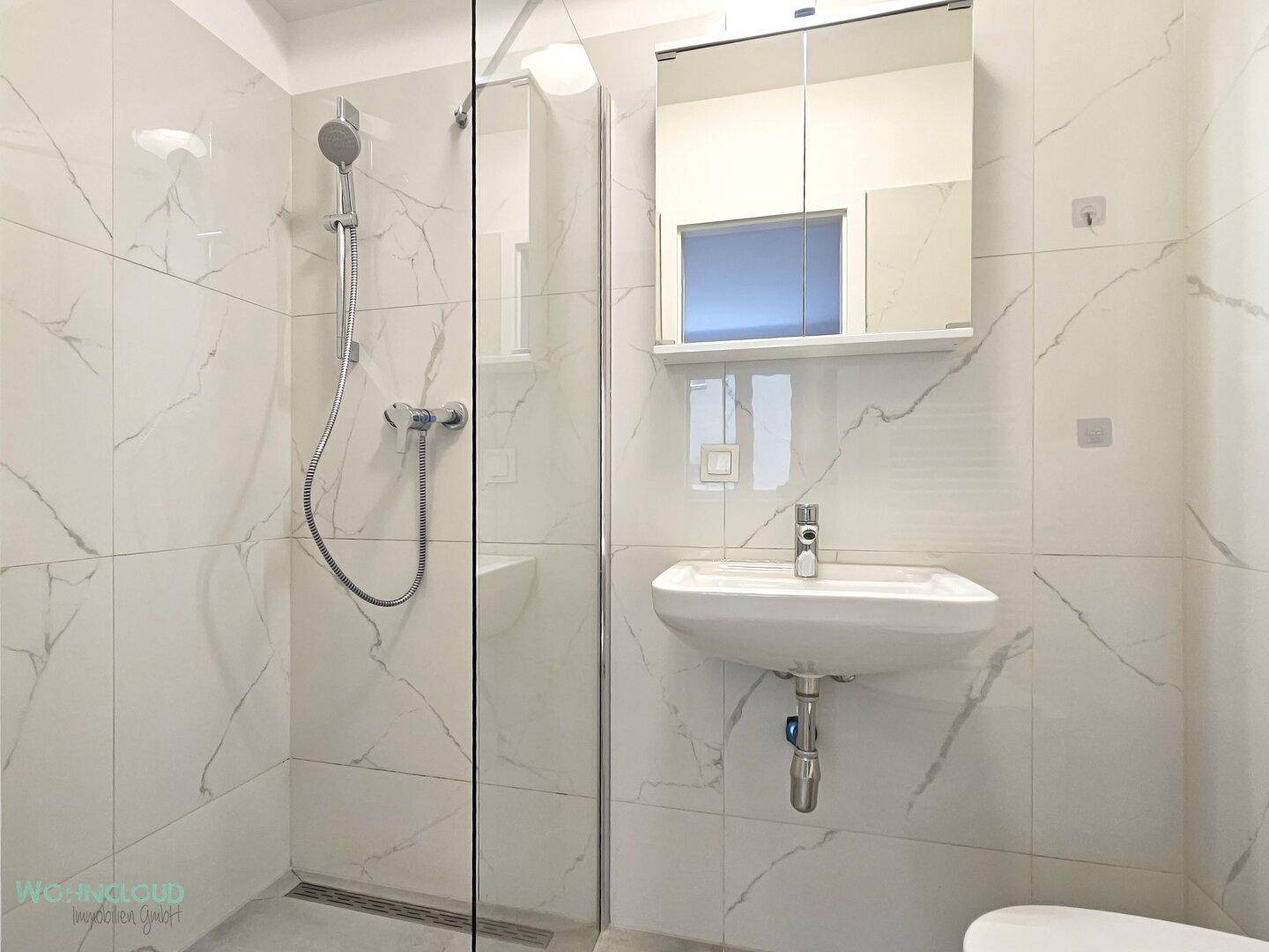 Modernes Badezimmer mit weißen Marmorfliesen, ebenerdiger Dusche und Waschtisch mit Spiegelschrank.