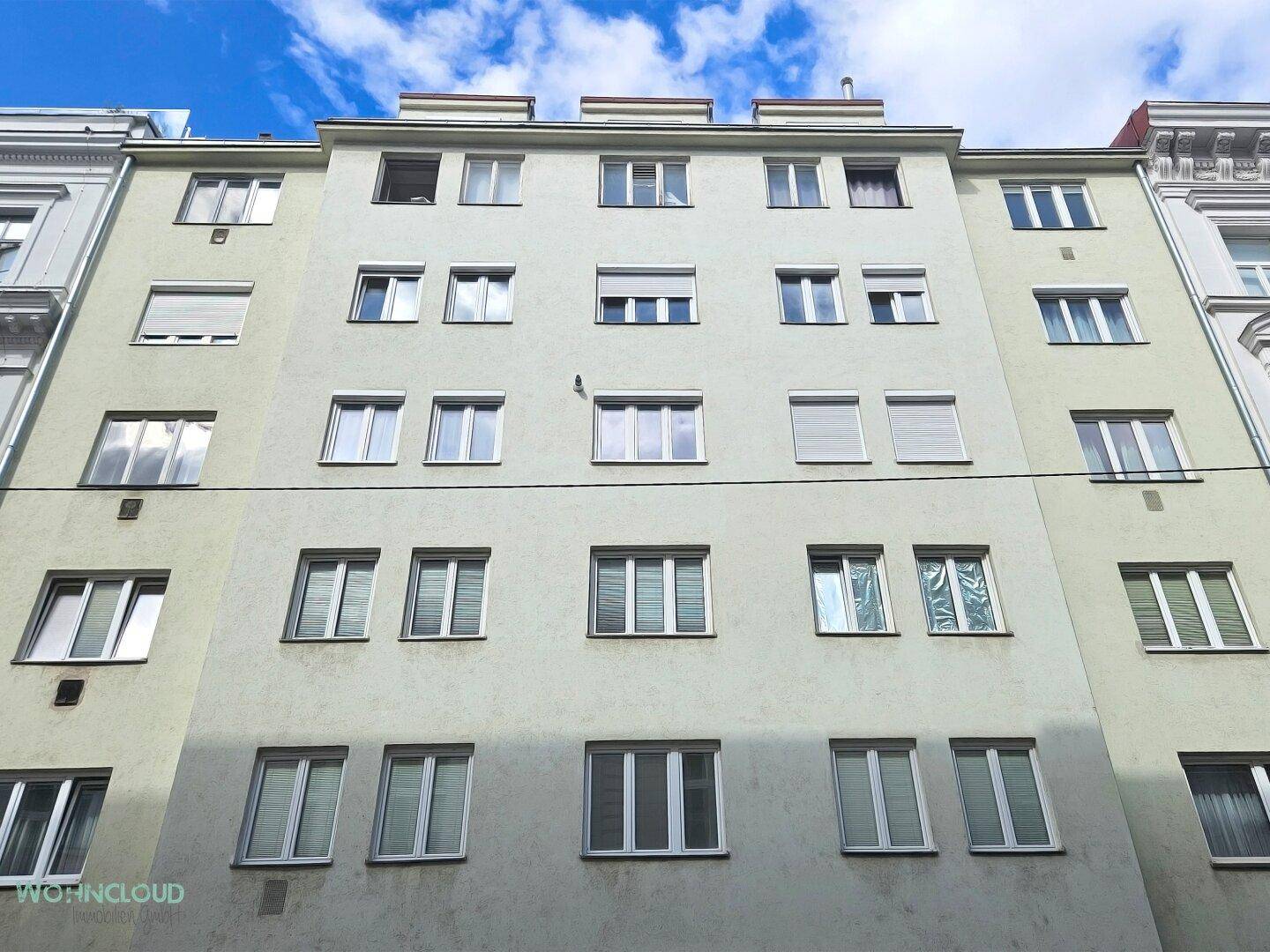 Fassade eines Mehrfamilienhauses mit zahlreichen Fenstern und Rollläden unter blauem Himmel.