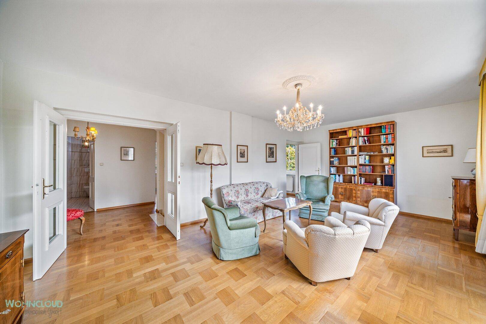 Geräumiges Wohnzimmer mit Parkettboden, Kronleuchter und einem großen Bücherregal.