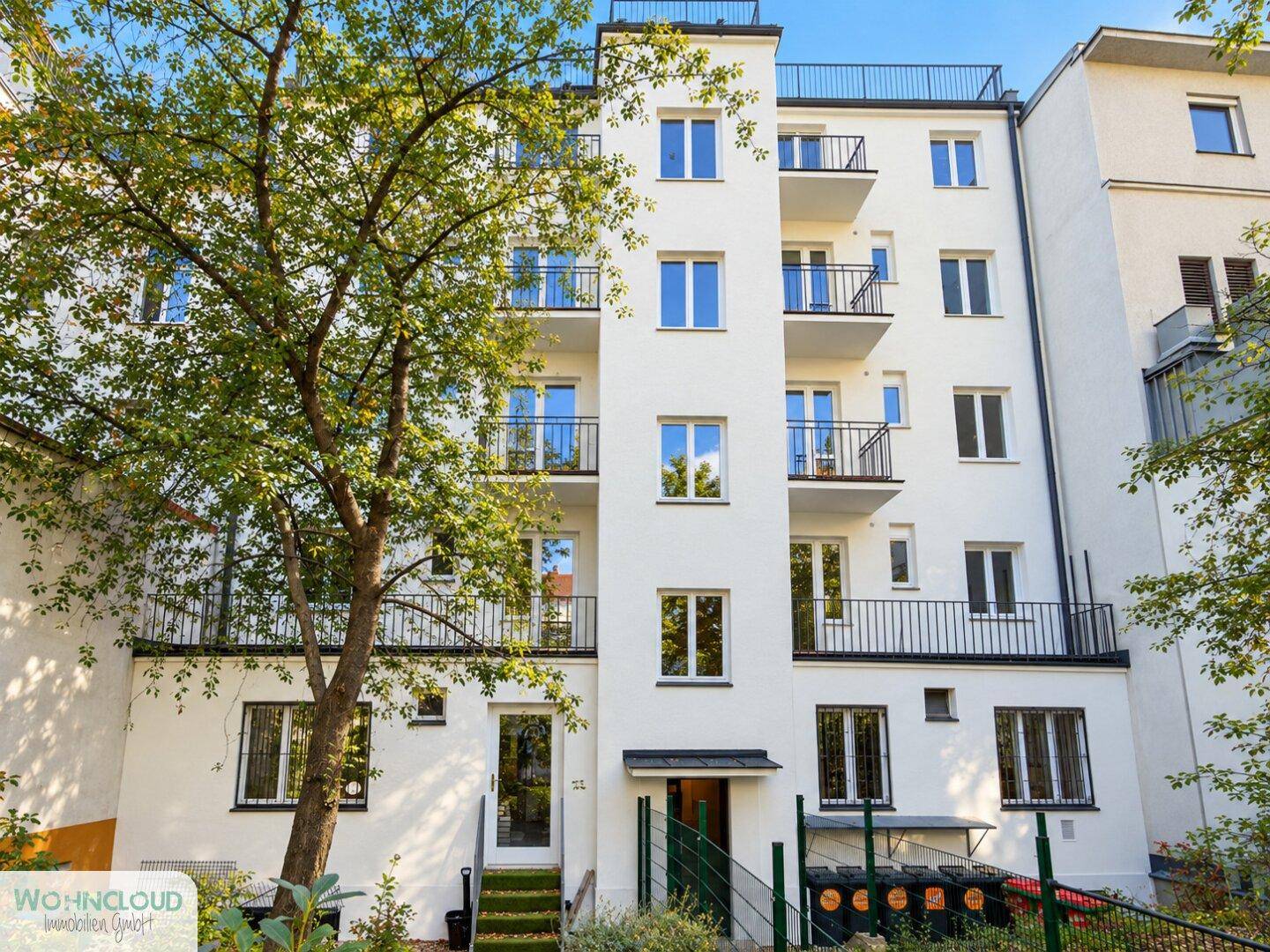 Moderne weiße Wohnhausfassade mit mehreren Balkonen und einem großen Baum im Vordergrund.