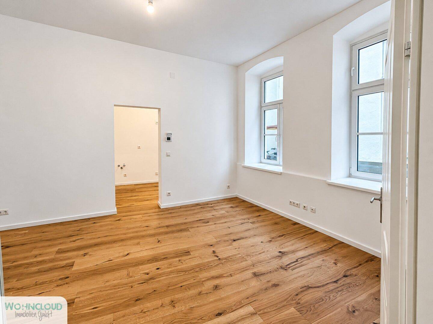 Geräumiges Zimmer mit hellem Holzboden und zwei Fenstern, ideal für Wohn- oder Schlafbereich.