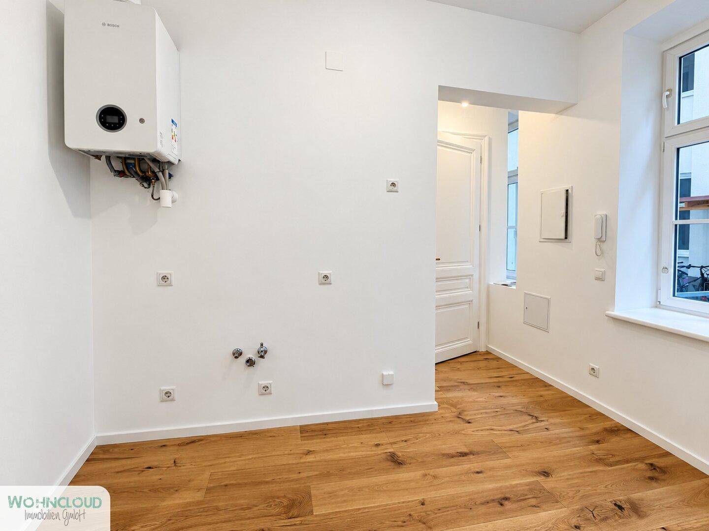 Raum mit Holzboden, Fenster und einem Warmwasserbereiter an der Wand, mit sichtbaren Anschlüssen.