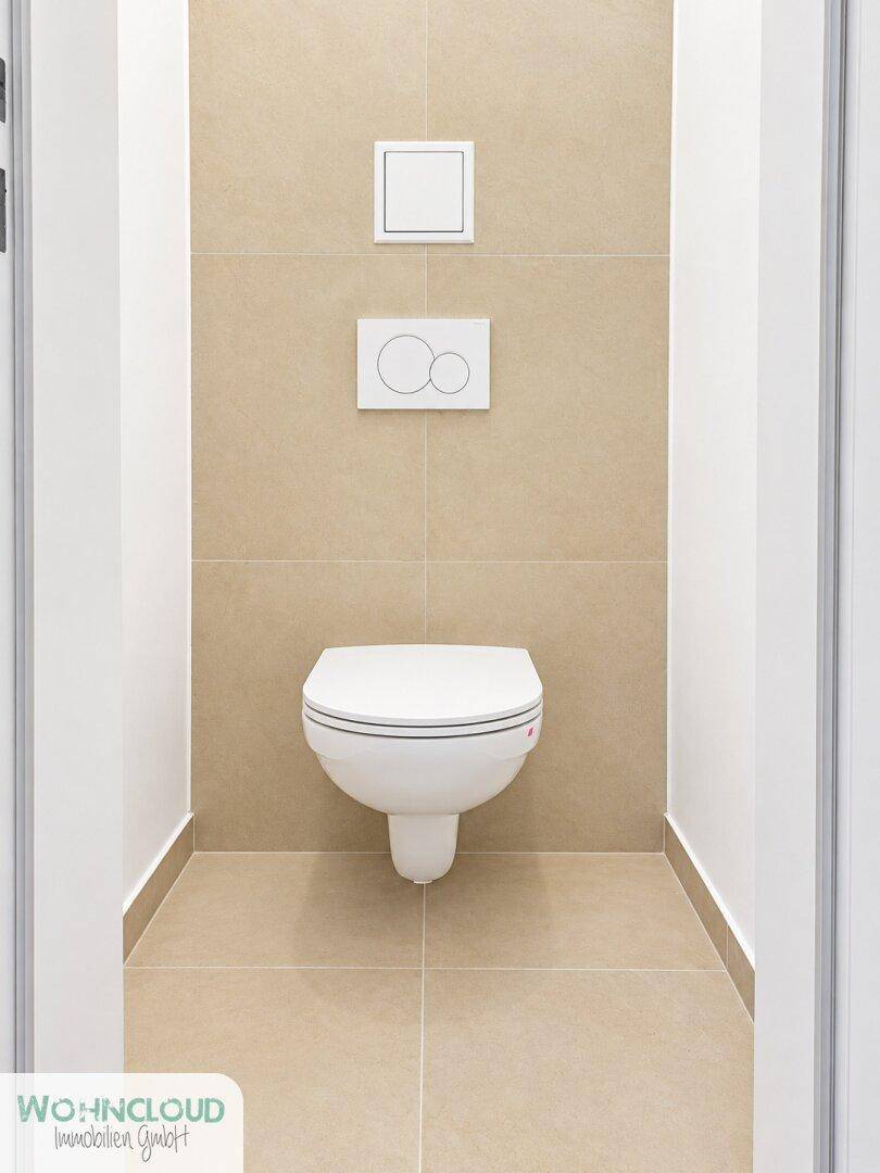 Separates WC mit wandhängender Toilette und beigefarbenen Fliesen an Wänden und Boden.