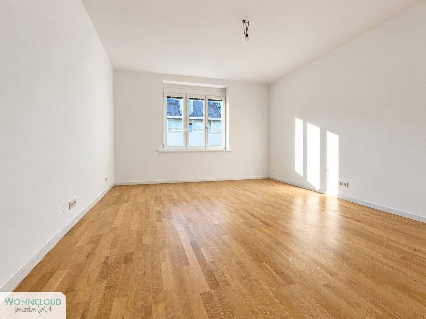 Helles, leeres Zimmer mit weiß gestrichenen Wänden und hellem Holzparkettboden.