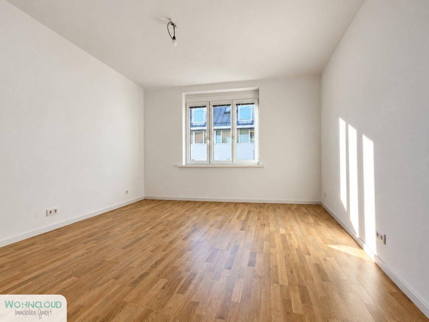 Helles, leeres Zimmer mit weiß gestrichenen Wänden und hellem Holzparkettboden.
