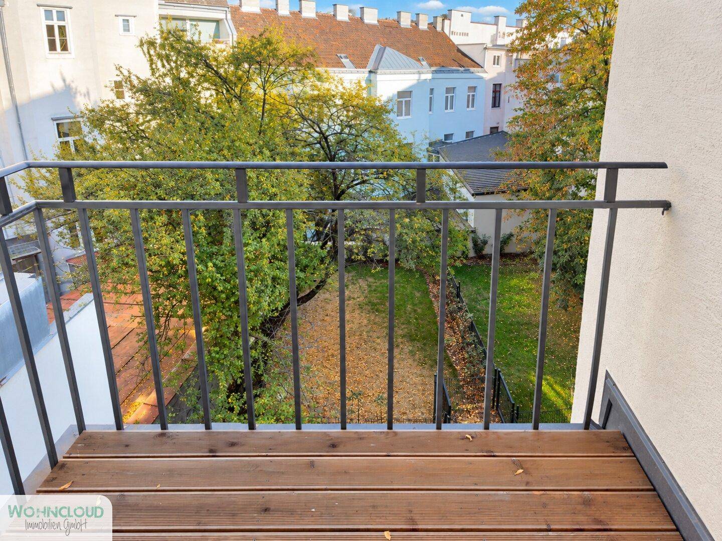 Kleiner Balkon mit Holzboden und Blick auf den grünen Innenhof mit Bäumen.
