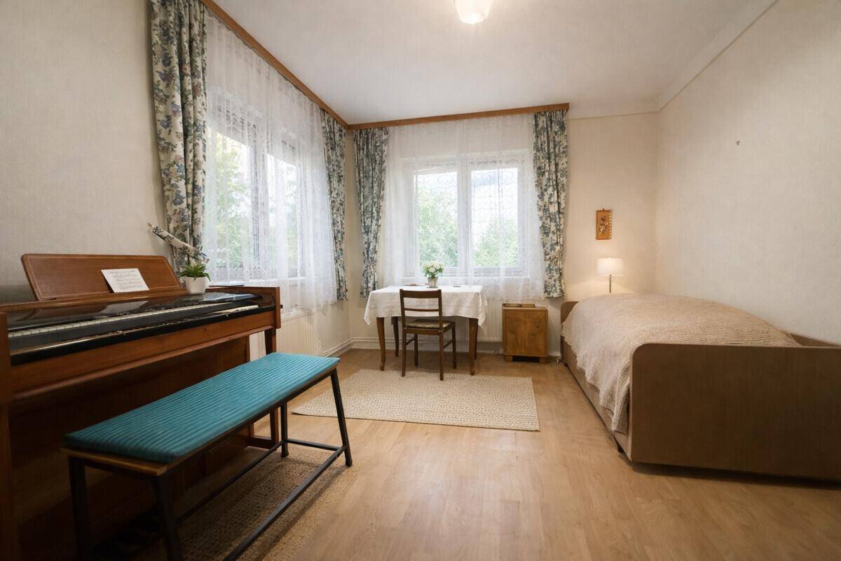 Helles Zimmer mit Einzelbett, Klavier und zwei Fenstern, die viel Tageslicht hereinlassen.