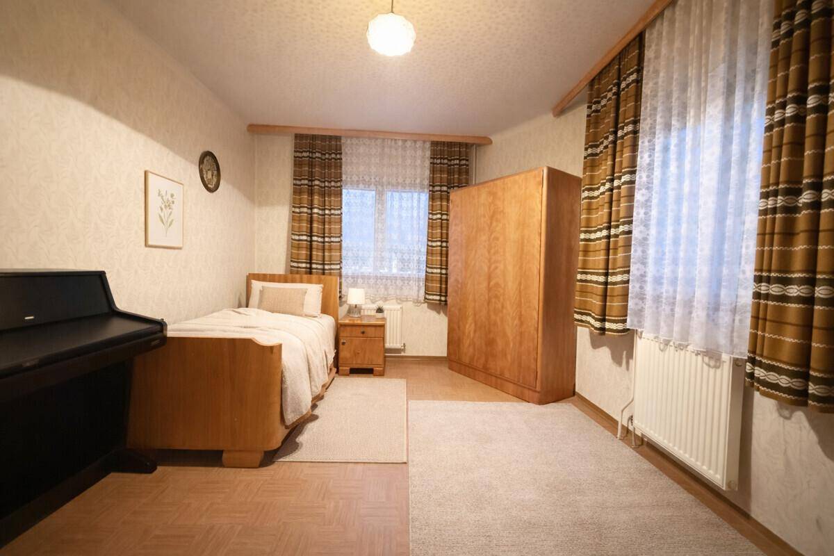 Gemütliches Schlafzimmer mit Einzelbett, Holzschrank und gemusterten Vorhängen an den Fenstern.