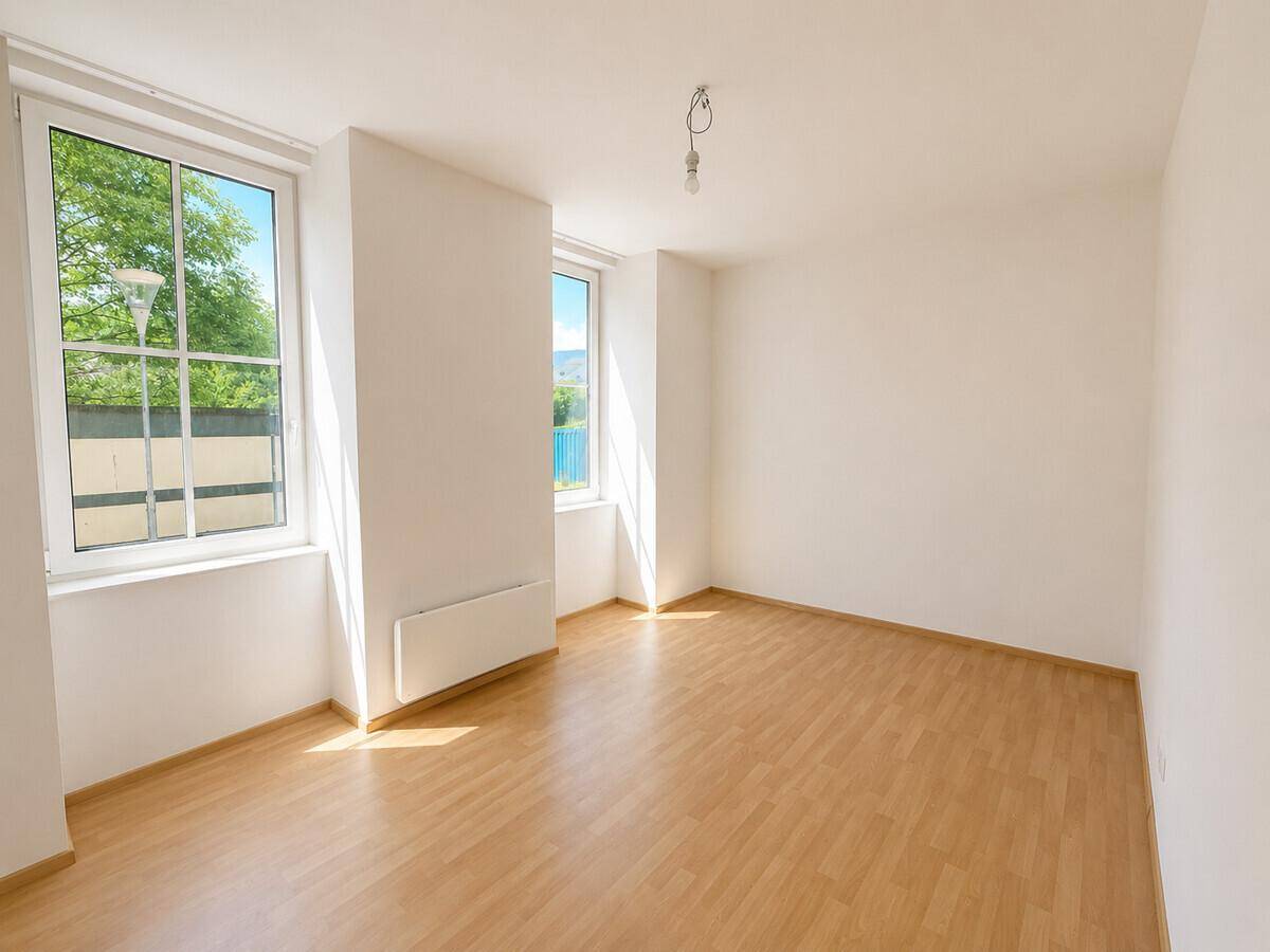 Helles, leeres Zimmer mit Laminatboden und großen Fenstern, die viel Tageslicht hereinlassen.