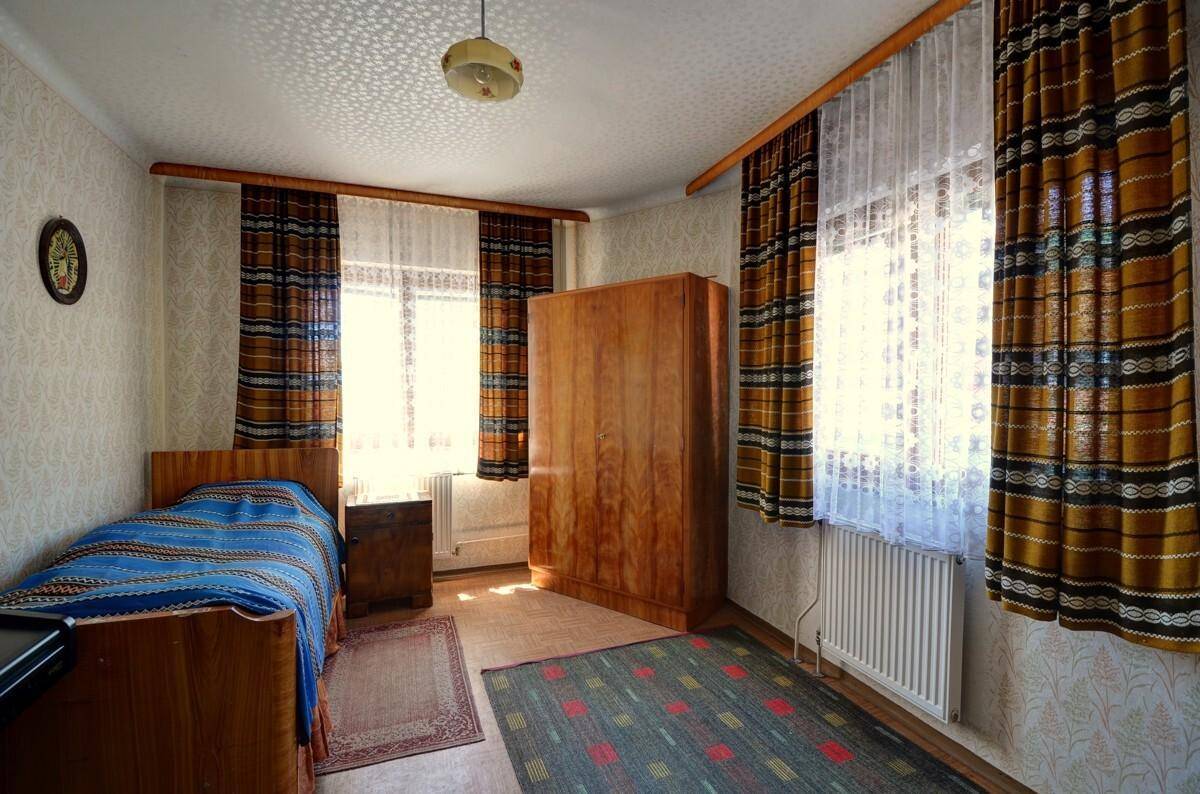 Schlafzimmer mit Einzelbett, Holzschrank und gemusterten Vorhängen an den Fenstern.