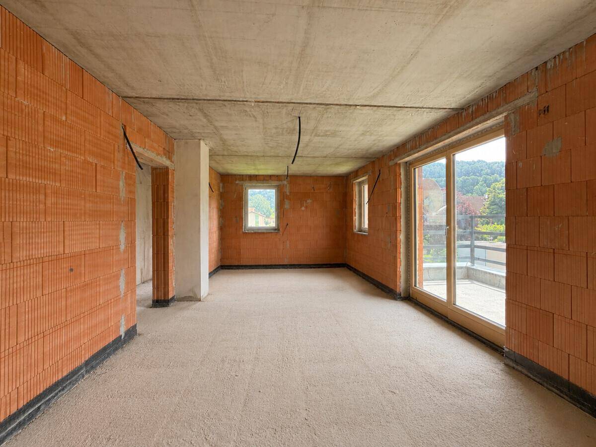Geräumiger Wohnbereich im Rohbau mit großen Fenstern und Zugang zum Balkon.