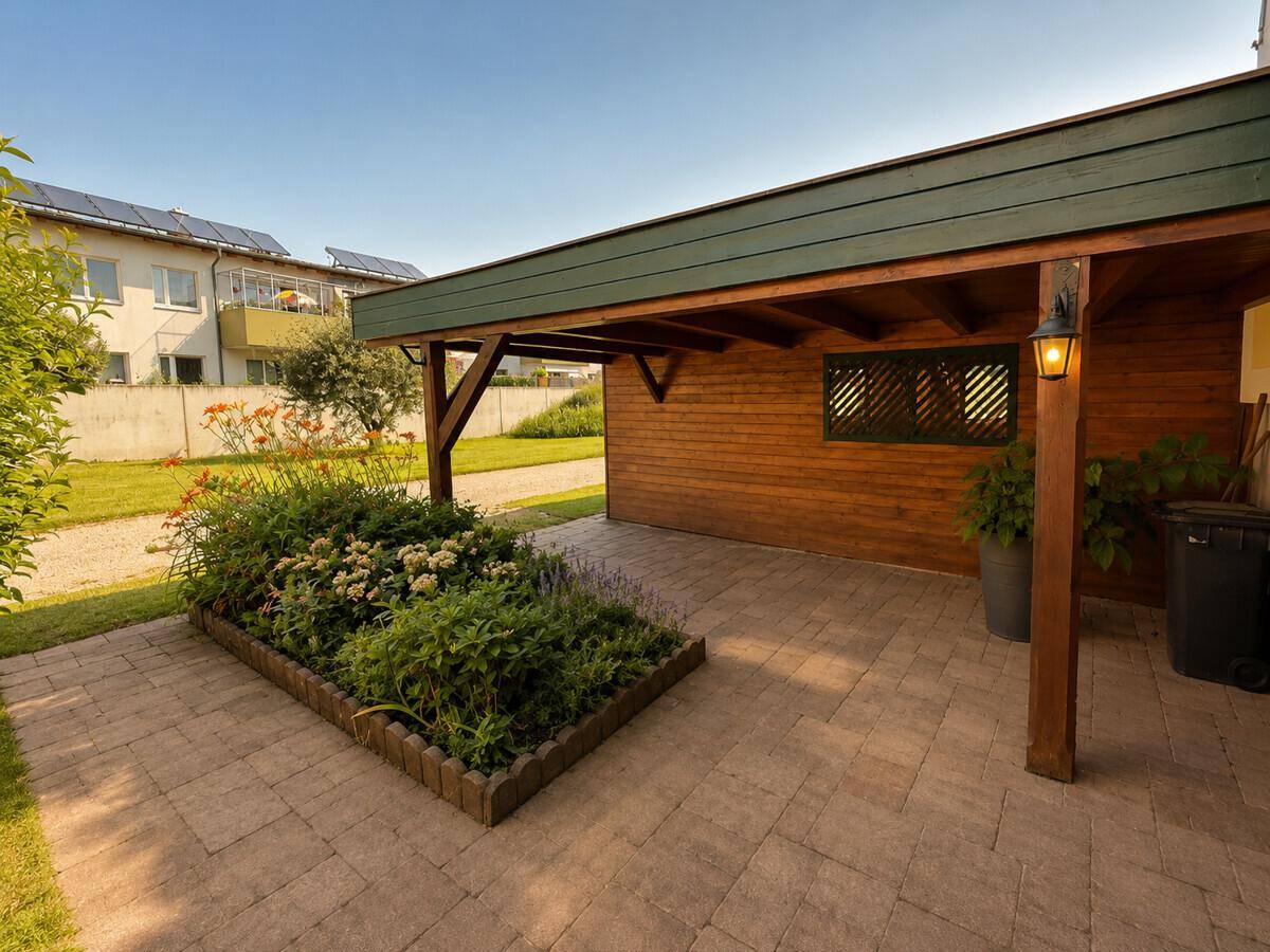 Überdachter Carport mit Holzbalken, gepflastertem Boden und Blumenbeet.