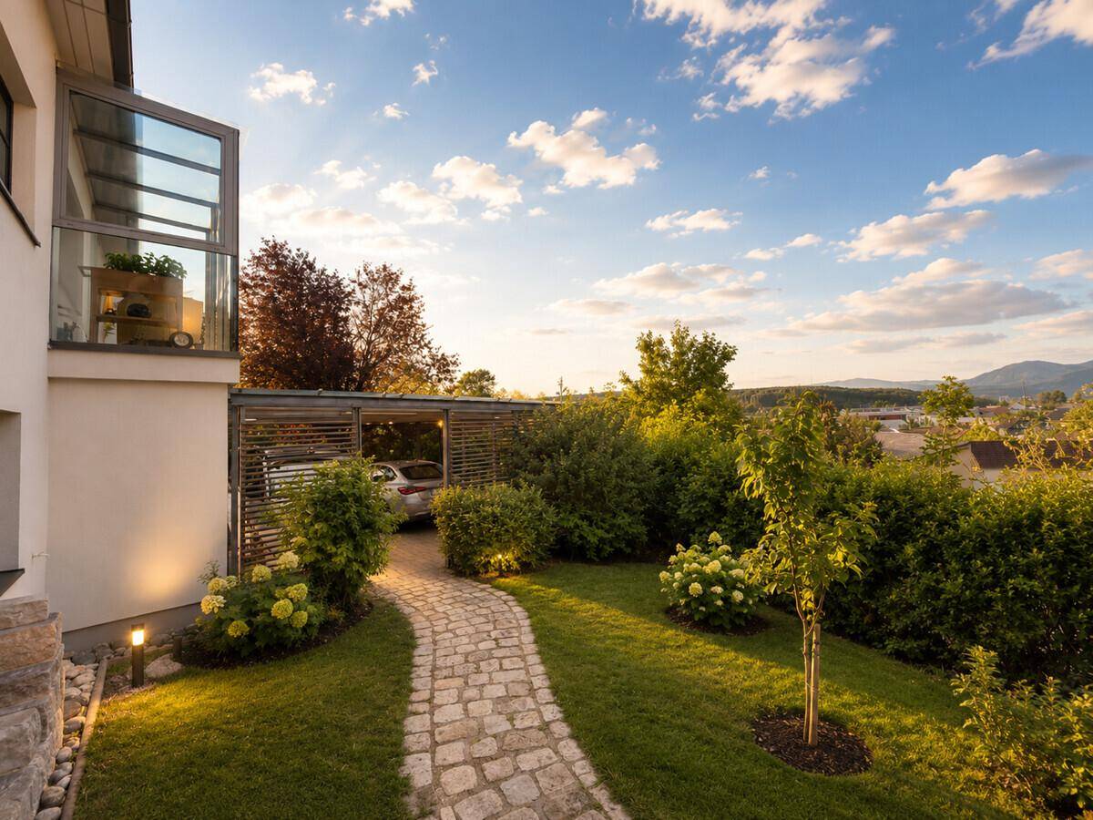 Gepflegter Garten mit Steinweg, üppiger Bepflanzung und Carport im Hintergrund bei Sonnenlicht.