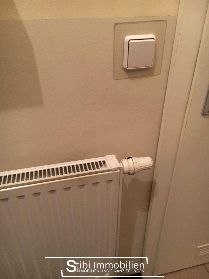 Weißer Heizkörper und Lichtschalter an einer hellen Wand neben einem Türrahmen in einem Innenraum.