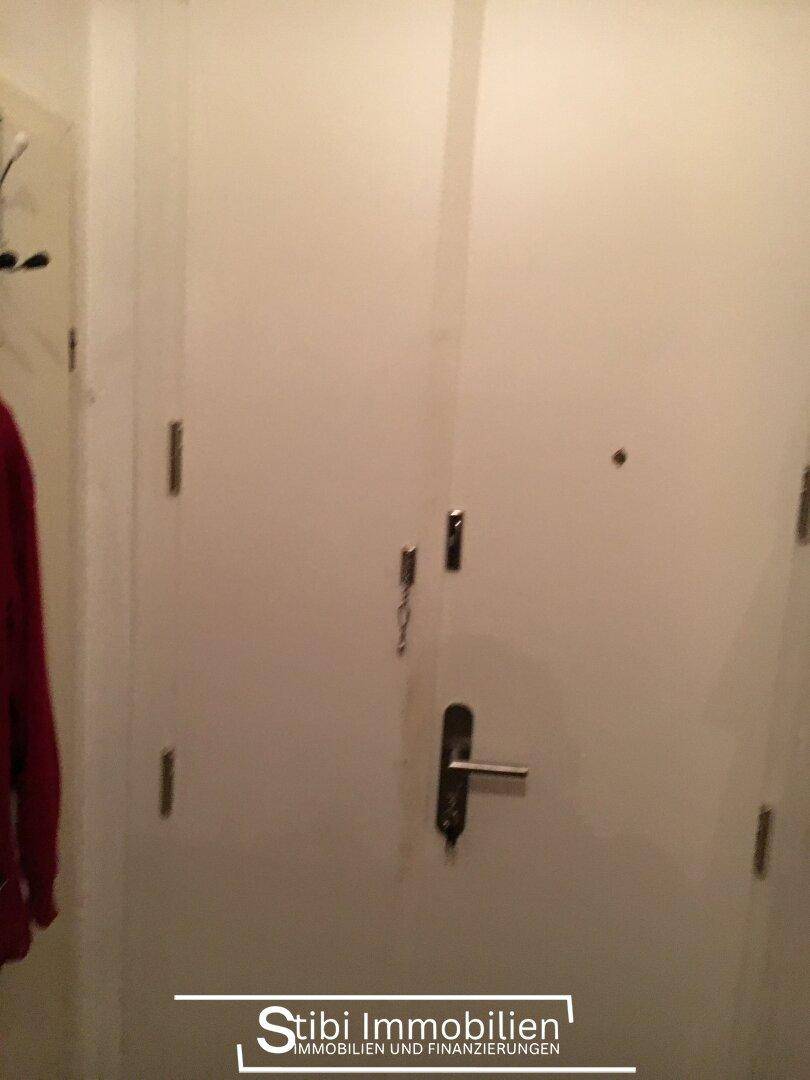 Weiße Eingangstür mit mehreren Schlössern und Griff, daneben hängt eine rote Jacke an einem Haken.