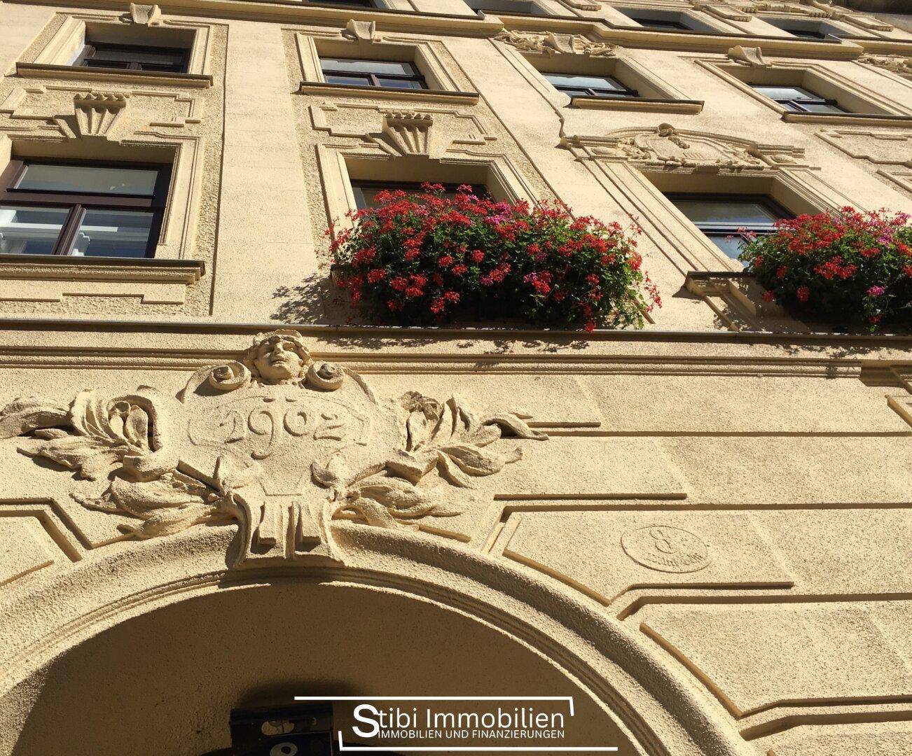 Historische Fassade eines Gebäudes von 1902 mit kunstvollen Verzierungen und Blumenkästen mit roten Blumen.