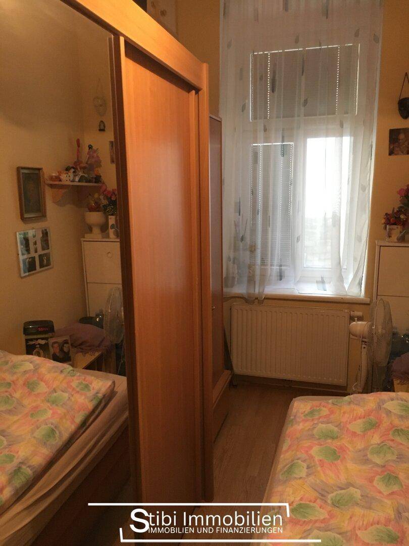 Helles Schlafzimmer mit zwei Betten, Holzschrank und Fenster mit Vorhängen und Heizkörper darunter.