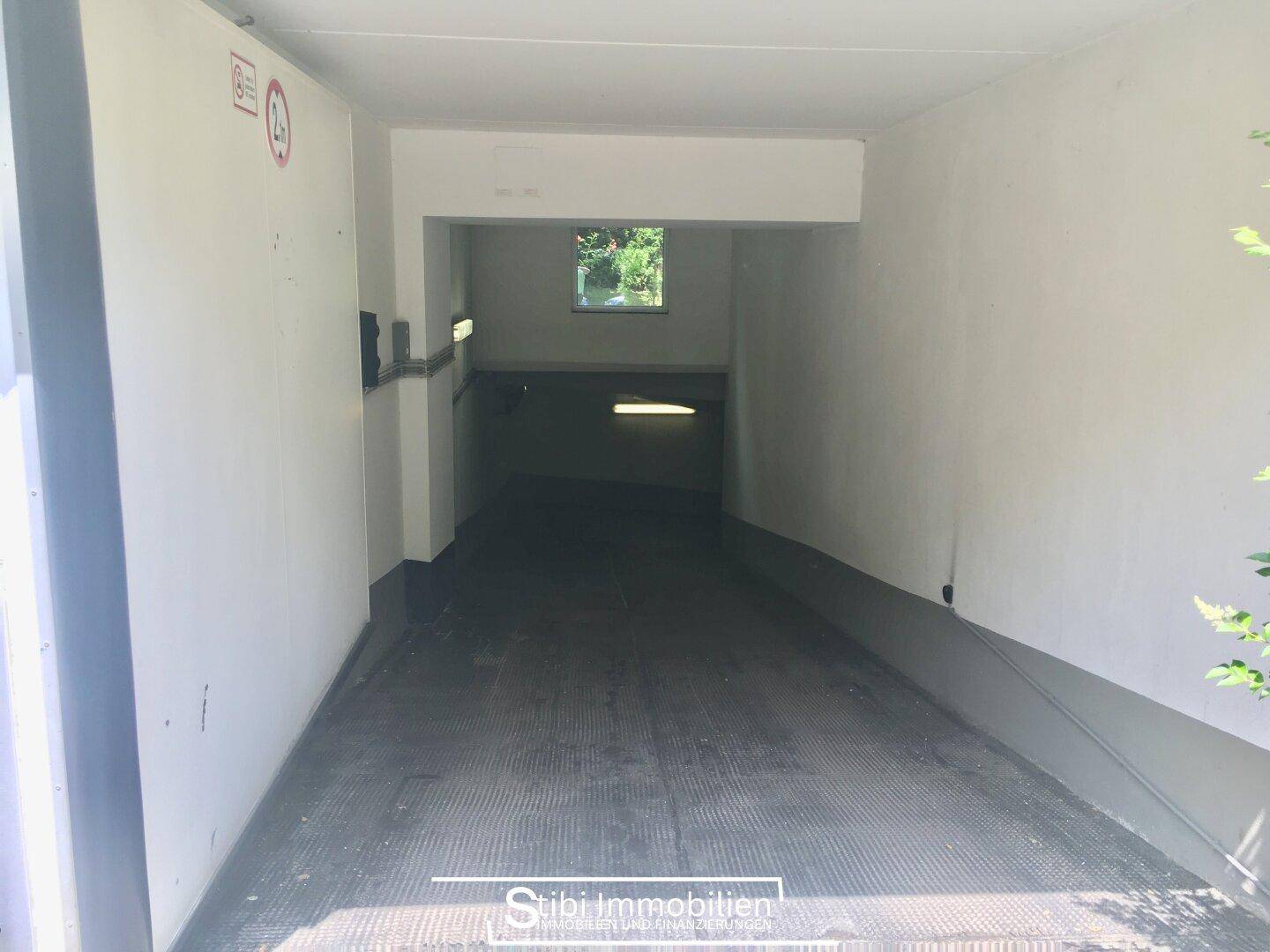 Steile Rampe führt in die Tiefgarage, mit hellen Wänden und einem Fenster, das Grünflächen zeigt.