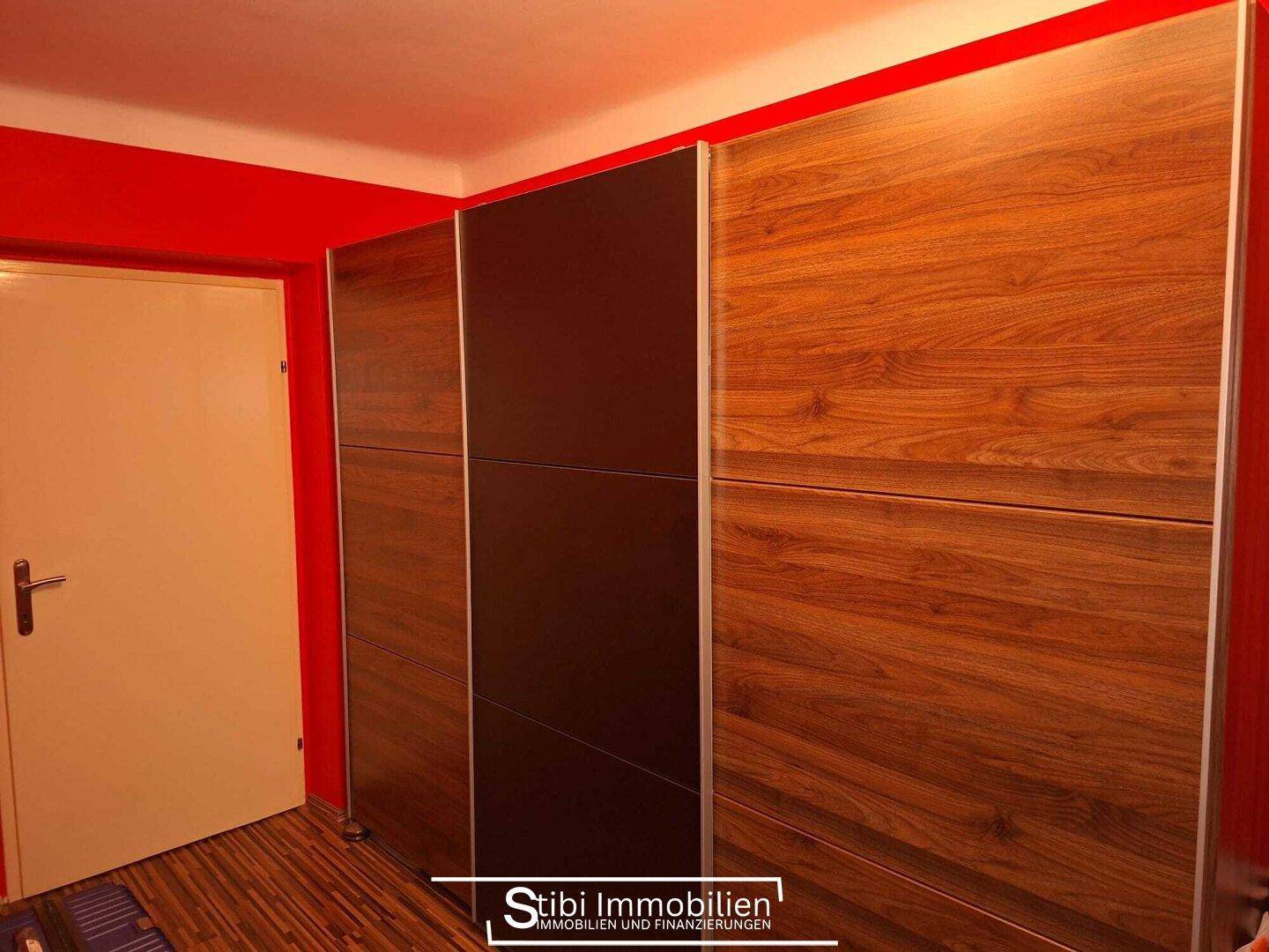 Schlafzimmer mit roter Wand, Holzschrank und dunklem Laminatboden.