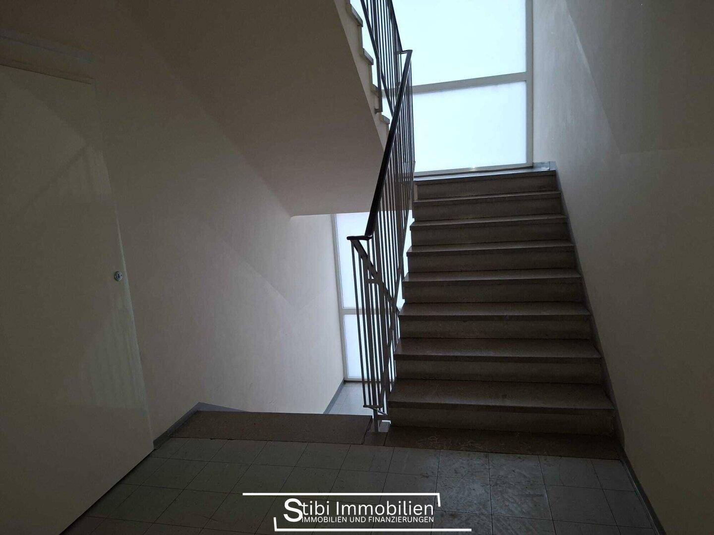 Treppenhaus mit Fliesenboden, Metallgeländer und Fenstern für Tageslicht.