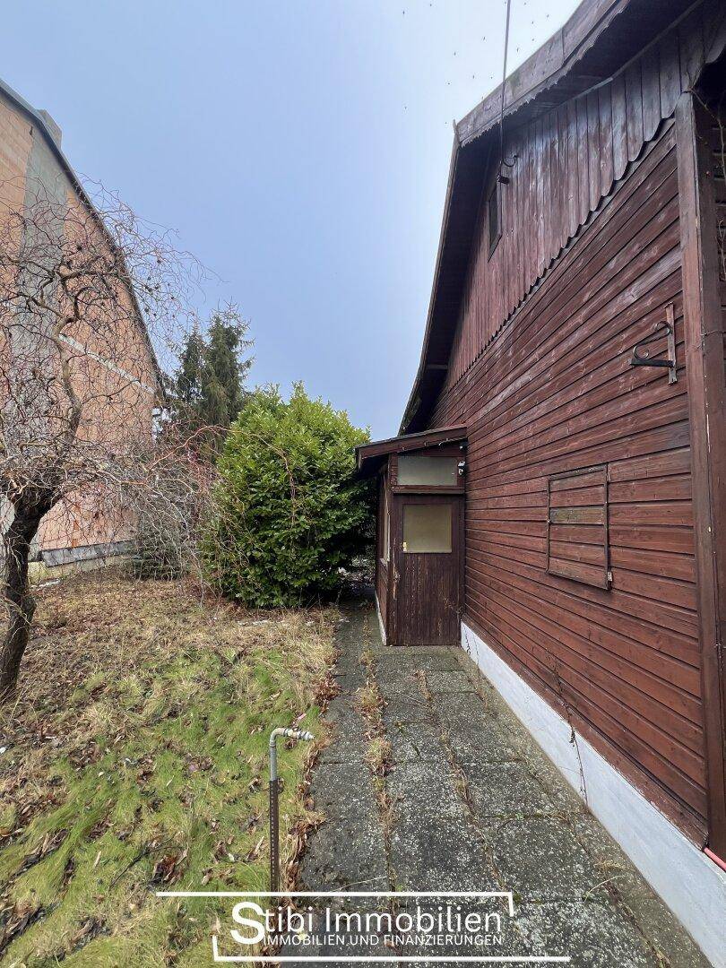Schmaler Weg entlang der Hauswand mit Holzeingang und verwildertem Gartenbereich.