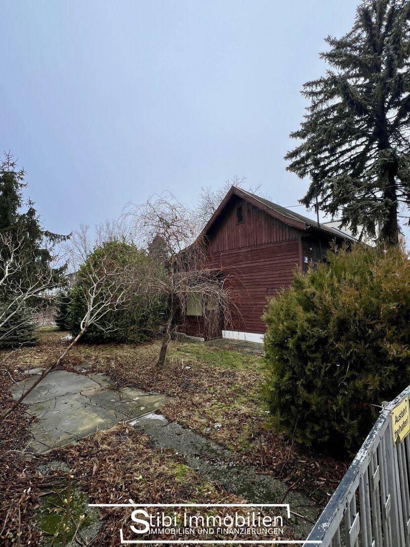 Altes Holzhaus mit Satteldach und verwildertem Garten, umgeben von Bäumen und einem Zaun.