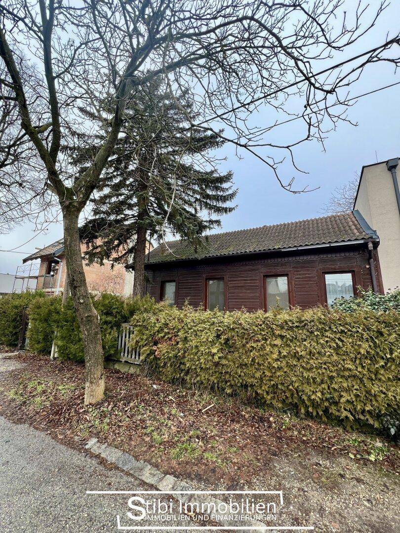 Vorderansicht des Holzhauses von der Straße aus, umgeben von einer hohen Hecke.