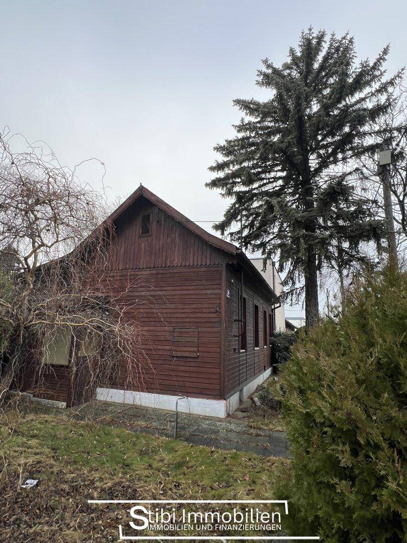 Ansicht des Holzhauses mit Satteldach und hohen Nadelbäumen im Gartenbereich.