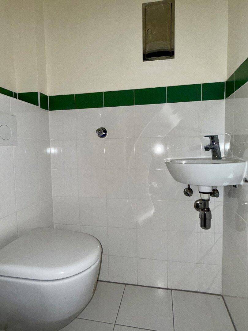 Gäste-WC mit Wand-WC, kleinem Waschbecken und weißen Fliesen mit grüner Bordüre.