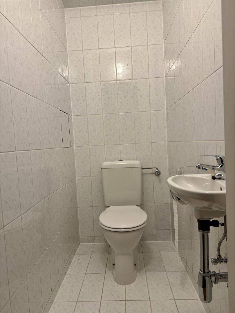 Separates WC mit weißen Fliesen, Toilette und kleinem Handwaschbecken.