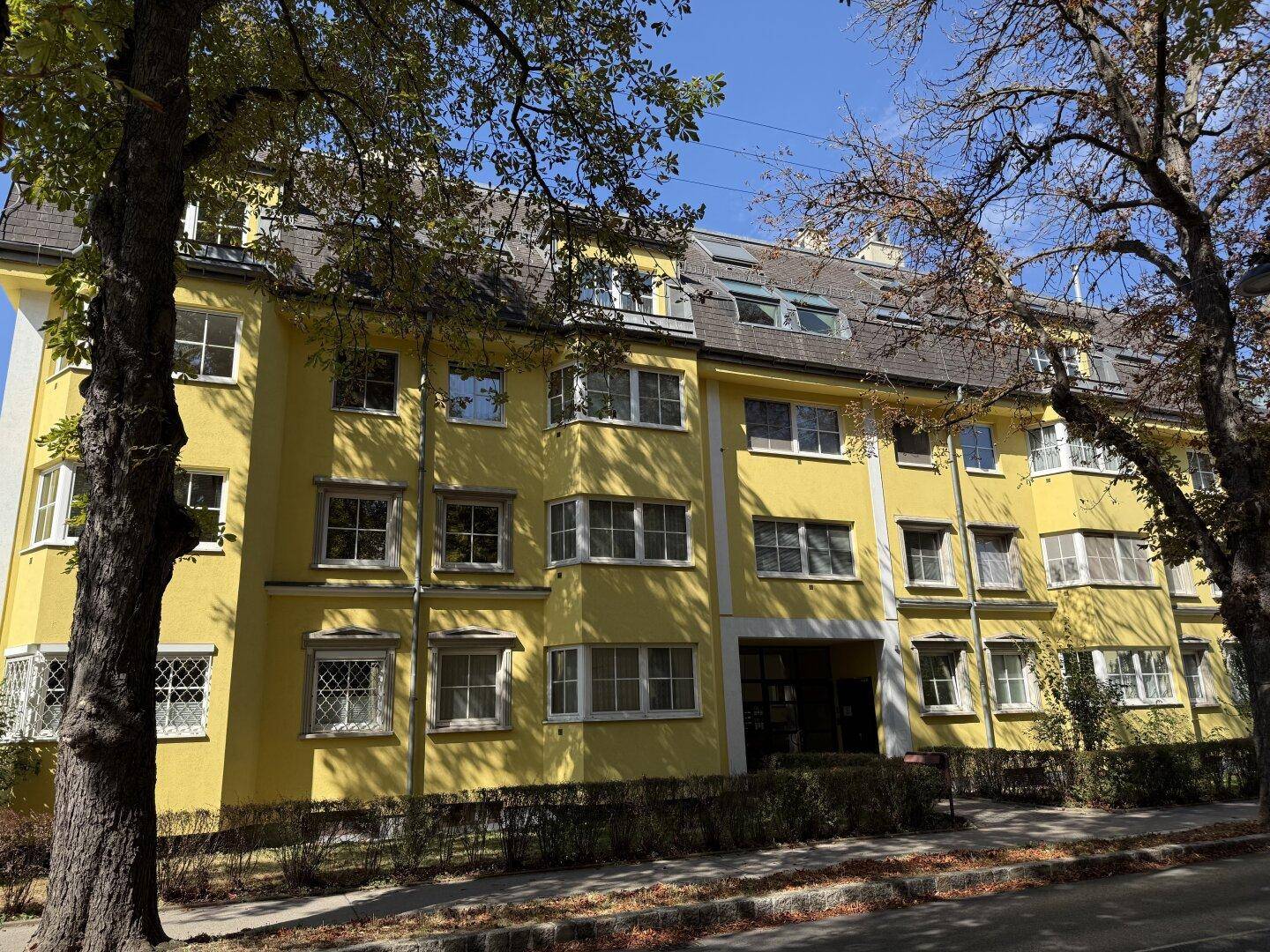 Attraktives gelbes Mehrfamilienhaus mit vielen Fenstern und altem Baumbestand davor.