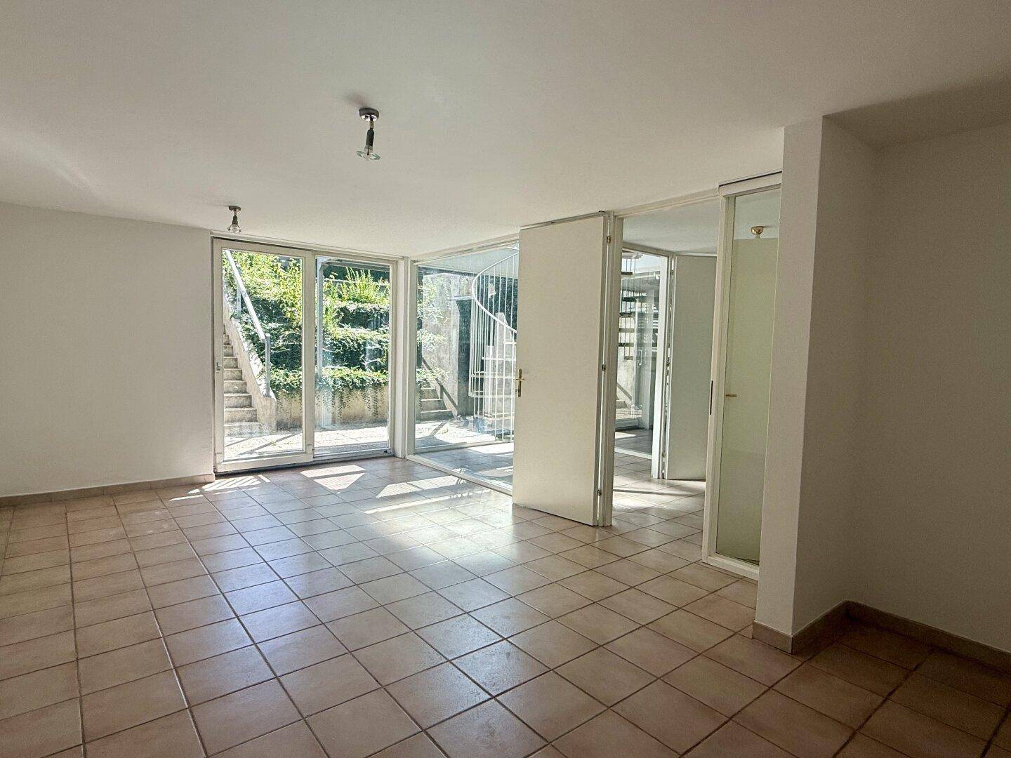 Geräumiger Wohnbereich mit Fliesenboden und viel Tageslicht durch große Fenster.