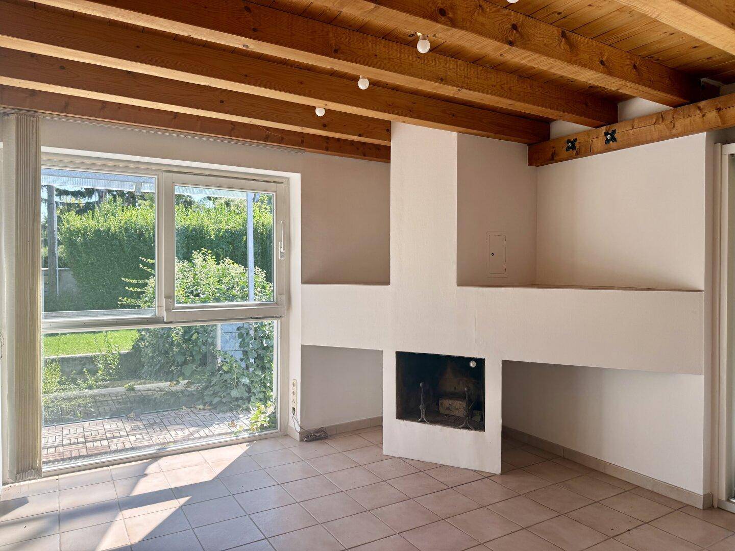 Helles Wohnzimmer mit Kamin, Fliesenboden und Blick ins Grüne durch große Fenster.