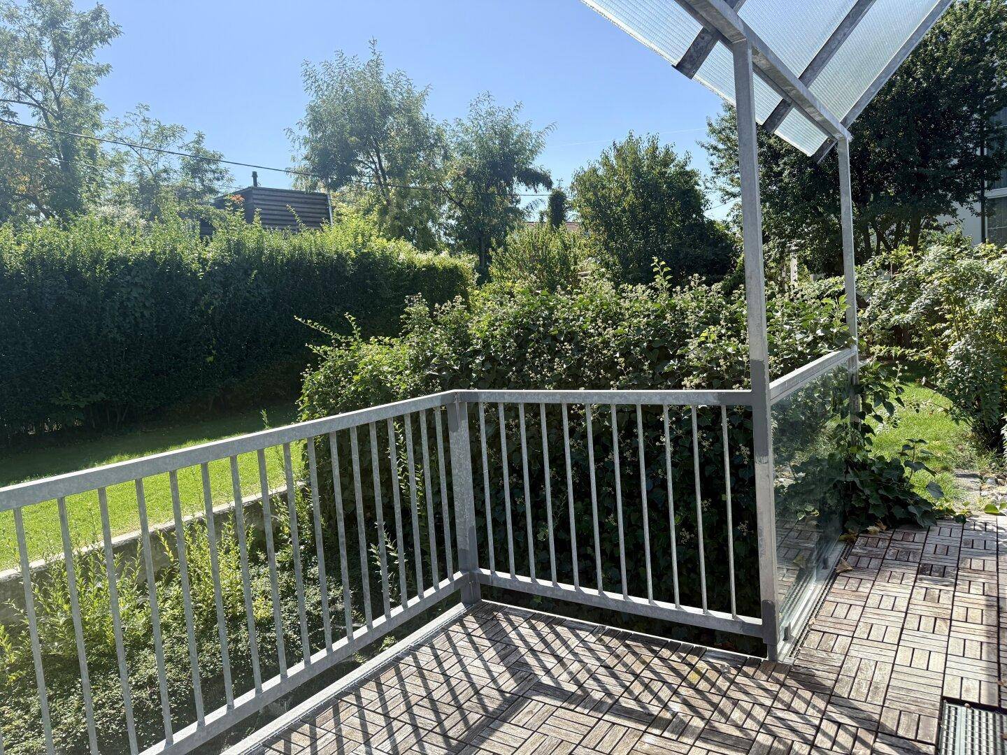 Überdachter Balkon mit Holzboden und Metallgeländer, umgeben von viel Grün.