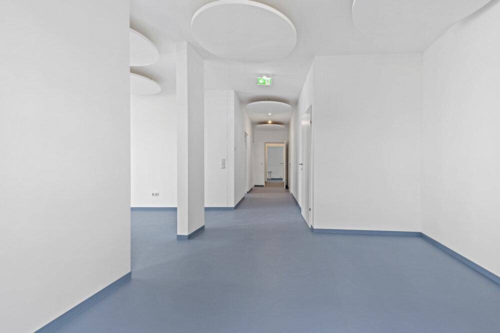 Langer, heller Flur mit weißen Wänden, blauem Boden und modernen Deckenelementen.